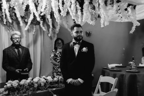 A formal gathering in a room decorated with cascading white flowers and draped fabric. A man in a suit stands beside a table adorned with floral arrangements, while another person in a patterned dress stands in the background, smiling. The ambiance is elegant and solemn.