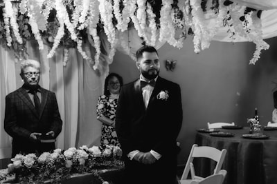 A formal gathering in a room decorated with cascading white flowers and draped fabric. A man in a suit stands beside a table adorned with floral arrangements, while another person in a patterned dress stands in the background, smiling. The ambiance is elegant and solemn.