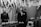 A formal gathering in a room decorated with cascading white flowers and draped fabric. A man in a suit stands beside a table adorned with floral arrangements, while another person in a patterned dress stands in the background, smiling. The ambiance is elegant and solemn.
