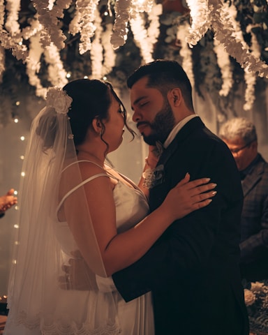 A bride and groom embracing during their wedding ceremony surrounded by cascading white flowers and soft, warm lighting that creates an intimate and romantic atmosphere.