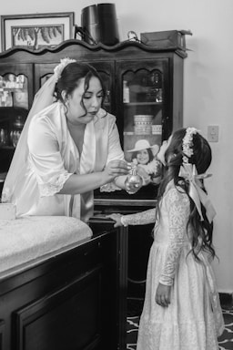 A woman wearing a bridal dress and veil is gently applying perfume to a young girl who is dressed in a lace outfit with floral headpieces. They are indoors, standing near a dark wooden cabinet with glass doors, containing various items. The scene appears calm and intimate, capturing a preparatory moment possibly for a wedding.