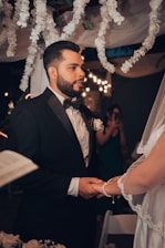 A man in a formal black suit with a bow tie is holding hands with a person wearing a white bridal gown and veil. They appear to be engaged in a wedding ceremony under a canopy decorated with white flowers. Soft, warm lighting and blurred background enhance the intimate setting.