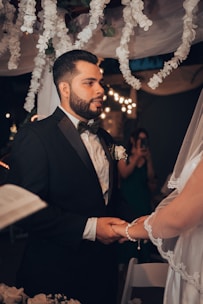 A man in a formal black suit with a bow tie is holding hands with a person wearing a white bridal gown and veil. They appear to be engaged in a wedding ceremony under a canopy decorated with white flowers. Soft, warm lighting and blurred background enhance the intimate setting.