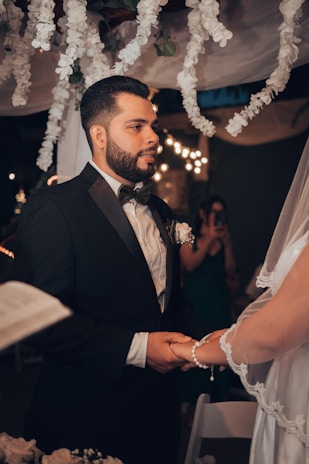 A man in a formal black suit with a bow tie is holding hands with a person wearing a white bridal gown and veil. They appear to be engaged in a wedding ceremony under a canopy decorated with white flowers. Soft, warm lighting and blurred background enhance the intimate setting.