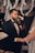 A man in a formal black suit with a bow tie is holding hands with a person wearing a white bridal gown and veil. They appear to be engaged in a wedding ceremony under a canopy decorated with white flowers. Soft, warm lighting and blurred background enhance the intimate setting.