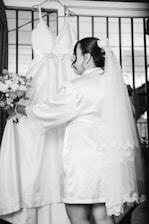 A woman wearing a bridal robe and veil is standing in front of a wedding dress hanging on a hanger. She holds a bouquet of flowers and appears to be preparing for a wedding ceremony.