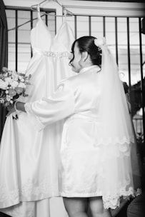 A woman wearing a bridal robe and veil is standing in front of a wedding dress hanging on a hanger. She holds a bouquet of flowers and appears to be preparing for a wedding ceremony.