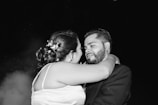 A black and white photograph depicts a couple embracing intimately, possibly during a special occasion. The woman, with floral decorations in her hair, faces the man, who is wearing a suit. They gaze at each other with affection.
