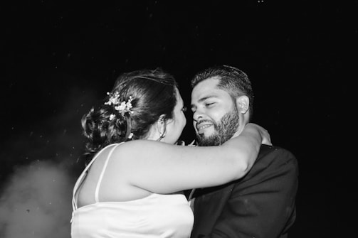A black and white photograph depicts a couple embracing intimately, possibly during a special occasion. The woman, with floral decorations in her hair, faces the man, who is wearing a suit. They gaze at each other with affection.