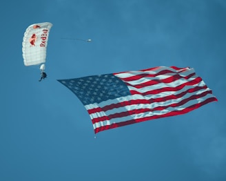 A skydiver descends with a white parachute, attached to which is a large, waving American flag. The sky is clear and blue, providing a striking backdrop to the scene.