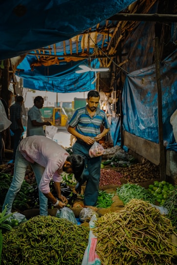 A bustling market scene with vendors and customers surrounded by piles of fresh vegetables. The area is covered by a blue tarp, creating a cozy atmosphere. Various vegetables are displayed in front of the vendors, including green beans, peas, and limes. Activity is evident as people are engaged in selecting and bagging produce.