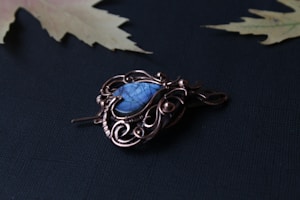 A copper wire-wrapped pendant with a blue stone as the centerpiece lies on a dark surface. The design is intricate, featuring swirls and loops in the wirework. In the background, there are two blurred leaves with a pale color, placed on an almost black fabric.