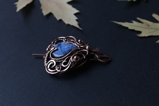 An ornate metal brooch with intricate swirling patterns and a central blue gemstone. The brooch rests on a dark textured fabric, surrounded by a few maple leaves lying nearby.