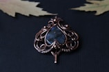 An intricate wire-wrapped pendant features a teardrop-shaped blue stone encased in ornate copper or bronze wirework. The background is a textured dark surface with two slightly blurred, pale yellow maple leaves visible in the corners.