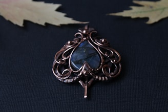 An intricate wire-wrapped pendant features a teardrop-shaped blue stone encased in ornate copper or bronze wirework. The background is a textured dark surface with two slightly blurred, pale yellow maple leaves visible in the corners.