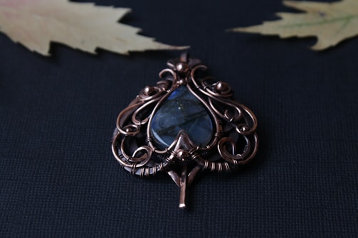 An intricate wire-wrapped pendant features a teardrop-shaped blue stone encased in ornate copper or bronze wirework. The background is a textured dark surface with two slightly blurred, pale yellow maple leaves visible in the corners.