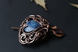 A beautifully crafted pendant with intricate copper wirework surrounding a blue gemstone, placed on a dark textured surface with a few leaves around it.