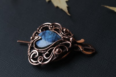 A beautifully crafted pendant with intricate copper wirework surrounding a blue gemstone, placed on a dark textured surface with a few leaves around it.