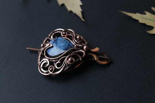 An intricately designed brooch featuring a blue gemstone at its center, encased in an ornate, swirling metal framework. The craftsmanship involves elegant curves and loops, suggesting an art nouveau style. The item is set against a dark, textured surface with a few light-colored, leaf-like elements nearby.