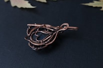 A piece of intricate wire-wrapped jewelry featuring a teardrop shape with detailed twisting and looping of copper-colored wires, set against a dark textured background. The design includes a smooth metal hook for attachment and a central glass or stone element.