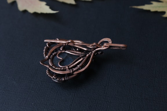 A piece of intricate wire-wrapped jewelry featuring a teardrop shape with detailed twisting and looping of copper-colored wires, set against a dark textured background. The design includes a smooth metal hook for attachment and a central glass or stone element.