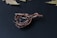 A piece of intricate wire-wrapped jewelry featuring a teardrop shape with detailed twisting and looping of copper-colored wires, set against a dark textured background. The design includes a smooth metal hook for attachment and a central glass or stone element.