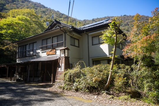 A traditional Japanese house surrounded by lush greenery, with a mountainous backdrop. The house features a wooden exterior with sliding windows and a gabled roof, blending harmoniously with the natural environment.