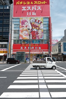A vibrant urban scene featuring a large building with colorful advertising graphics of animated characters on the facade. The building has signage in Japanese and advertises a pachinko and slot venue. At the foreground, a small white truck is driving across a crosswalk on a clear day. Adjacent buildings and a blue sky are also visible.