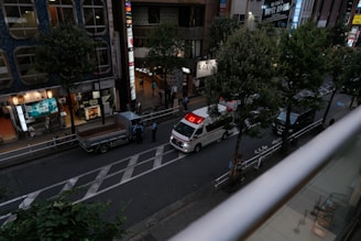 A street scene from an elevated perspective, showing an ambulance with its red lights illuminated parked on the road. Nearby, a small truck is stationed with a few people standing nearby, possibly police officers. The street is lined with trees and bordered by buildings with bright signs and lit storefronts, suggesting an urban environment, possibly in a commercial district.