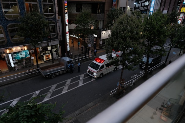 A street scene from an elevated perspective, showing an ambulance with its red lights illuminated parked on the road. Nearby, a small truck is stationed with a few people standing nearby, possibly police officers. The street is lined with trees and bordered by buildings with bright signs and lit storefronts, suggesting an urban environment, possibly in a commercial district.