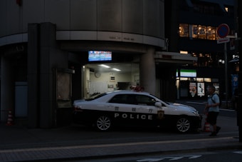 A police car is parked outside a police box or station at night. The surroundings are dimly lit with some artificial lighting coming from the station and nearby buildings. A person is walking past the police car on the sidewalk.
