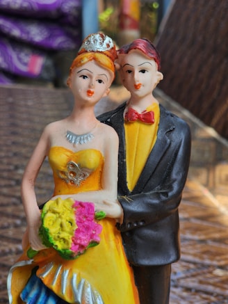 A figurine of a couple dressed in formal attire, with the female figure wearing a yellow dress and holding a bouquet of flowers, while the male figure is wearing a black suit with a yellow shirt and a red bow tie. They stand close together on a textured surface with a blurry background that includes hints of purple and blue.