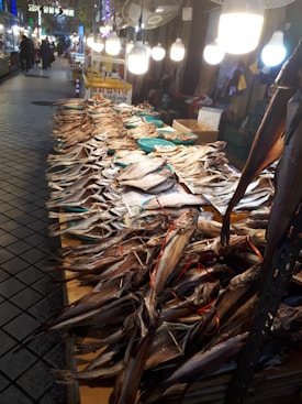 A market scene showcasing an array of dried fish laid out on a table under bright overhead lighting. The market is bustling with activity in the background, featuring vibrant signs and vendors. The fish are stacked in rows, some tied with red string, creating a textured and organized pattern.