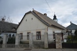 A traditional, rustic house with a gabled roof, featuring three windows framed in dark wood. It is surrounded by a stone fence with metal gates. In the background, a church steeple with a cross is visible, adding an element of cultural heritage. The sky is overcast, contributing to a calm, serene atmosphere.