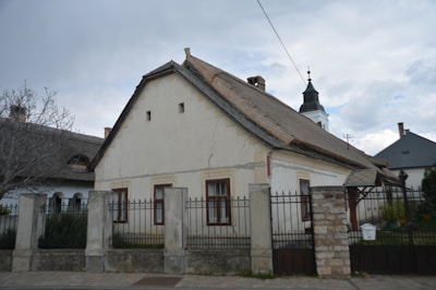 A traditional, rustic house with a gabled roof, featuring three windows framed in dark wood. It is surrounded by a stone fence with metal gates. In the background, a church steeple with a cross is visible, adding an element of cultural heritage. The sky is overcast, contributing to a calm, serene atmosphere.