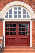 A double wooden door painted in red with glass panels, set in a brick wall with an arched transom window above. The bricks are reddish-orange, and there is a short staircase leading up to the door with terracotta tiles. White stone framing is present around the window.