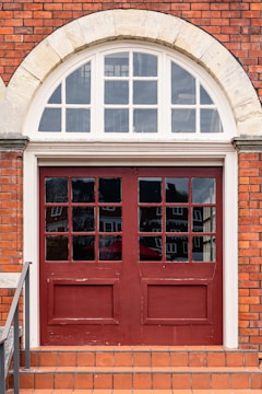 A double wooden door painted in red with glass panels, set in a brick wall with an arched transom window above. The bricks are reddish-orange, and there is a short staircase leading up to the door with terracotta tiles. White stone framing is present around the window.