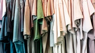 A collection of colorful t-shirts hanging closely together, arranged in a row. The shirts display a variety of shades, including blues, greens, and neutral tones. The material appears casual and lightweight, suitable for everyday wear.