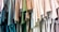 A collection of colorful t-shirts hanging closely together, arranged in a row. The shirts display a variety of shades, including blues, greens, and neutral tones. The material appears casual and lightweight, suitable for everyday wear.