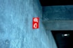 A red fire extinguisher sign is mounted on a textured concrete wall inside a building, with a modern design. The ceiling appears to have a dark panel and a recessed light fixture.