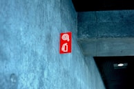 A red fire extinguisher sign is mounted on a textured concrete wall inside a building, with a modern design. The ceiling appears to have a dark panel and a recessed light fixture.