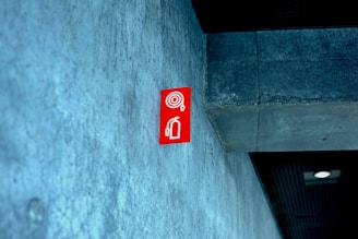 A red fire extinguisher sign is mounted on a textured concrete wall inside a building, with a modern design. The ceiling appears to have a dark panel and a recessed light fixture.