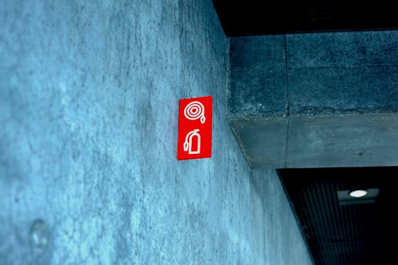 A red fire extinguisher sign is mounted on a textured concrete wall inside a building, with a modern design. The ceiling appears to have a dark panel and a recessed light fixture.