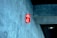 A red fire extinguisher sign is mounted on a textured concrete wall inside a building, with a modern design. The ceiling appears to have a dark panel and a recessed light fixture.