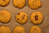 Nine cookies are arranged on a sheet of brown parchment paper. Each cookie has a golden-brown color with visible bits of chocolate or dried fruit embedded in the dough.