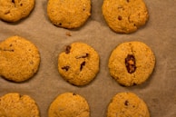 Nine cookies are arranged on a sheet of brown parchment paper. Each cookie has a golden-brown color with visible bits of chocolate or dried fruit embedded in the dough.