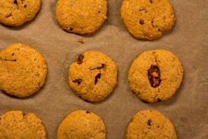 Nine cookies are arranged on a sheet of brown parchment paper. Each cookie has a golden-brown color with visible bits of chocolate or dried fruit embedded in the dough.