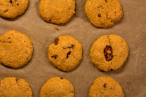 Nine cookies are arranged on a sheet of brown parchment paper. Each cookie has a golden-brown color with visible bits of chocolate or dried fruit embedded in the dough.