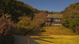 A large, traditional-style house is situated on a grassy hill surrounded by lush trees. The building features multiple windows and a steeply pitched roof. A path leads up to the house, bordered by trees and shrubs. The scene is bathed in warm sunlight, casting soft shadows across the landscape.