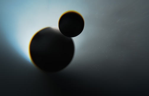 Two overlapping dark circular shapes are set against a gradient background transitioning from light blue to dark. The composition creates an impression of celestial bodies or planets, with subtle lighting effects highlighting their silhouettes.